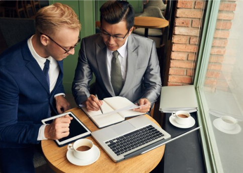 Two men in business attire sit at a round table in a cafe. Their intense focus suggests they are conducting a risk assessment for vendors.