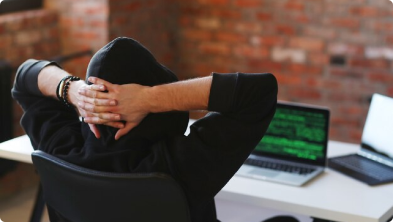 Person in a black hoodie sitting at a desk, leaning back with hands behind head, looking at a laptop