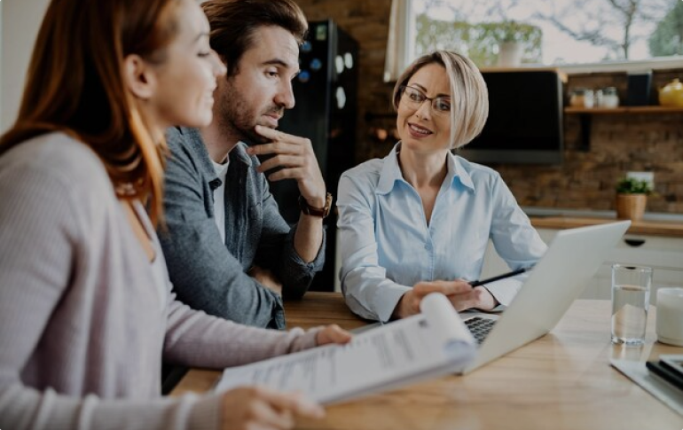 Three people sit around a table with documents and a laptop. The person on the right, appears to be explaining something about real estate development to the other two.