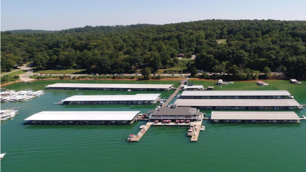 Aerial view of a marina with multiple covered docks and boats on a green-hued lake, surrounded by dense forested hills under a clear sky, providing clients with an idyllic retreat.