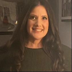 A woman with long dark hair and a green sweater exudes leadership as she smiles while standing indoors.