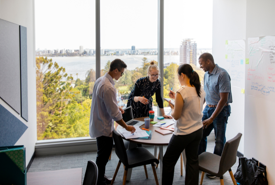 Four people stand around a table in a modern office with large windows overlooking a city skyline and river.