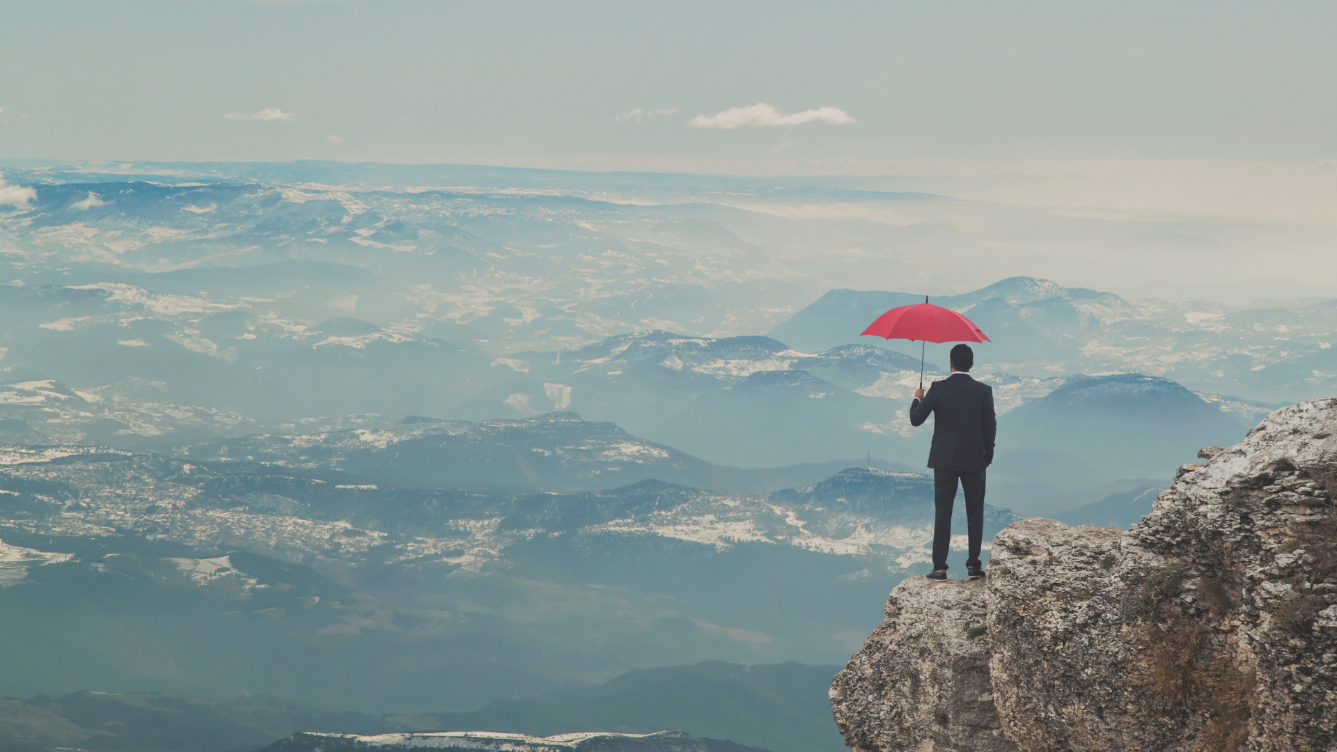 A man in a suit stands on a rocky cliff holding a red umbrella, overlooking a vast mountain landscape