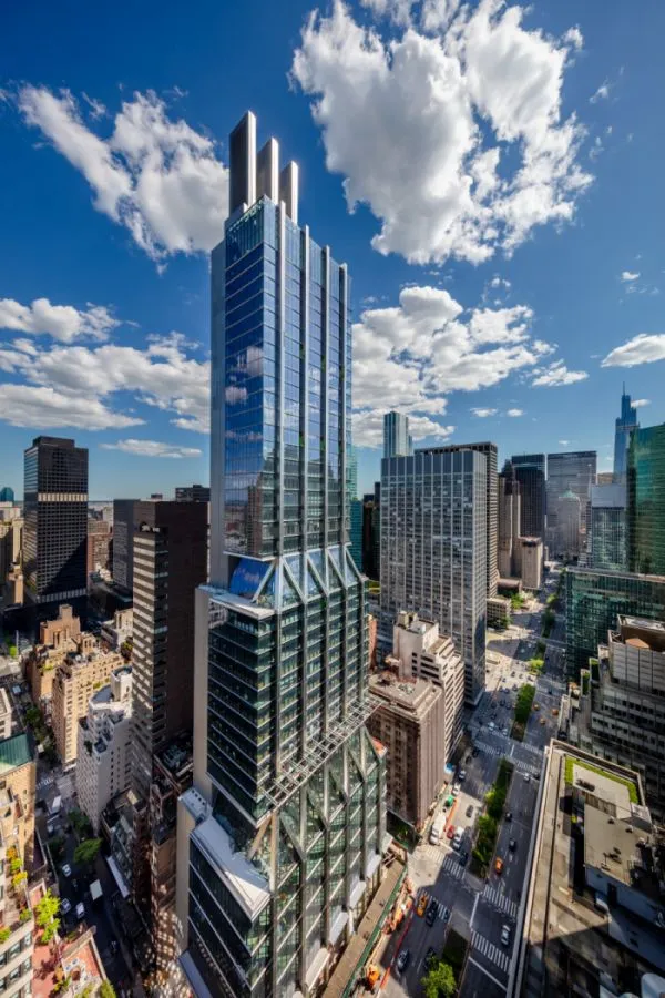 An aerial view of a modern skyscraper with a glass exterior surrounded by other tall buildings under a partly cloudy sky in a bustling cityscape, capturing the vibrant hub where clients meet and news unfolds.