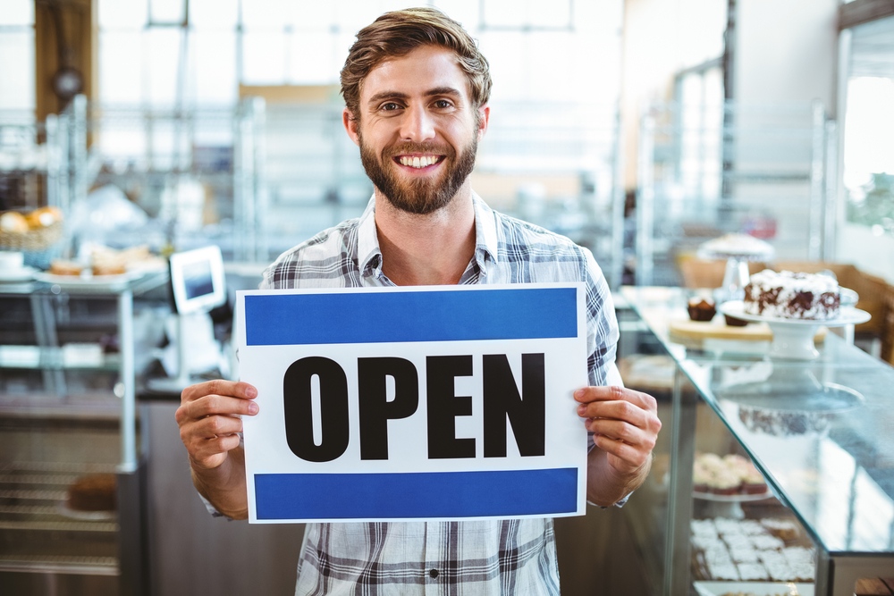 A man in a plaid shirt holds an "OPEN" sign inside a bakery, with shelves of baked goods in the background.