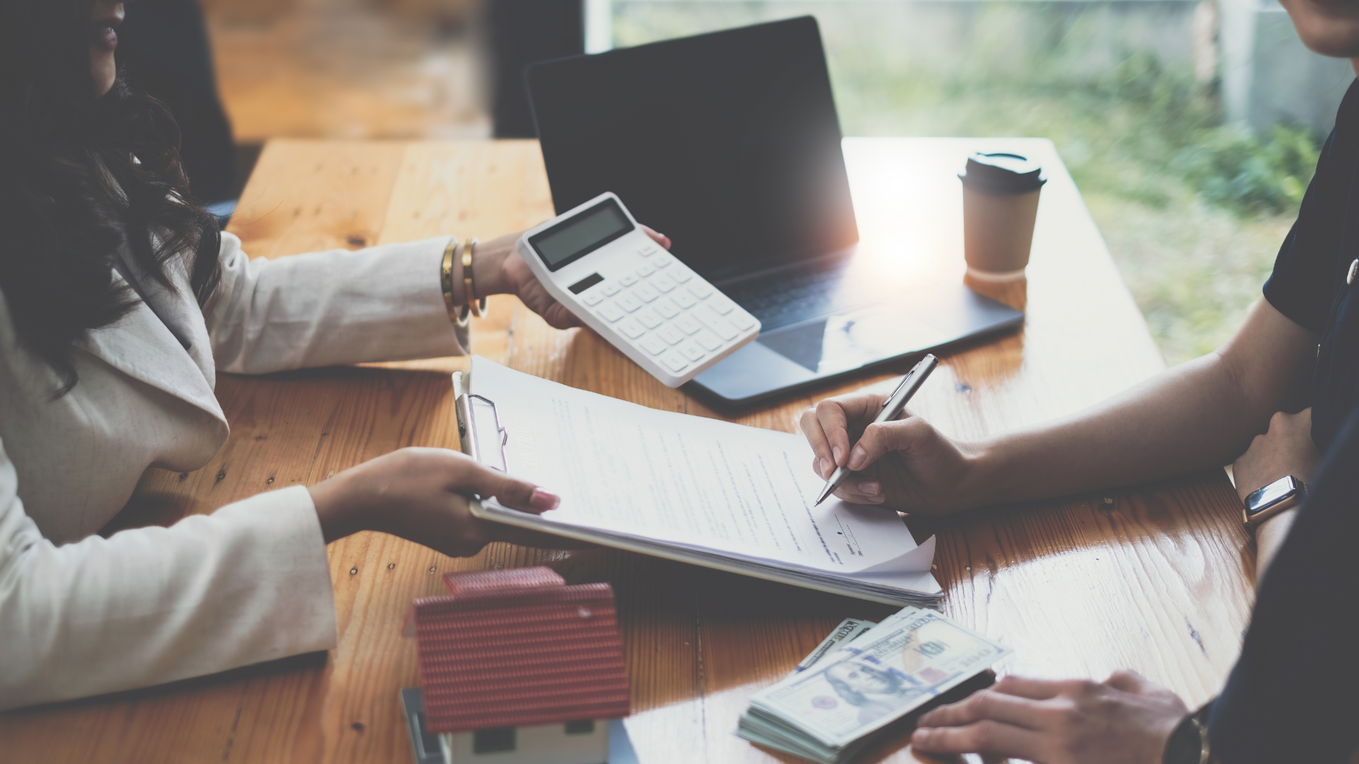 Two people sit at a wooden table. One person holds a clipboard with documents related to claims management.