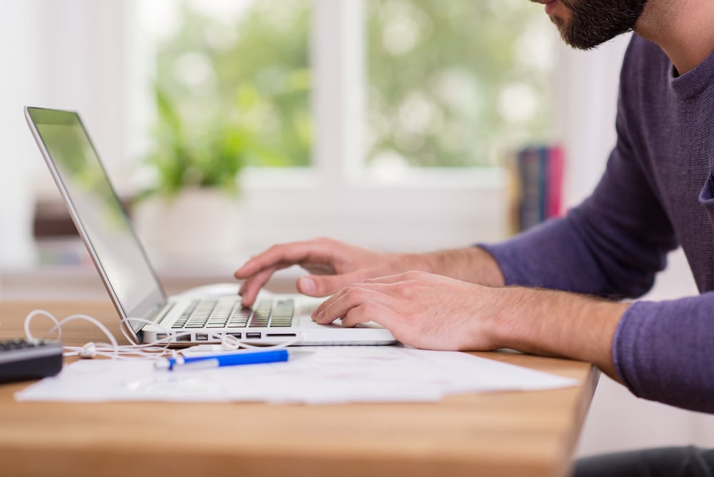 A risk management consultant typing on a laptop at a desk, with papers, a pen, earphones, and a plant in the background.