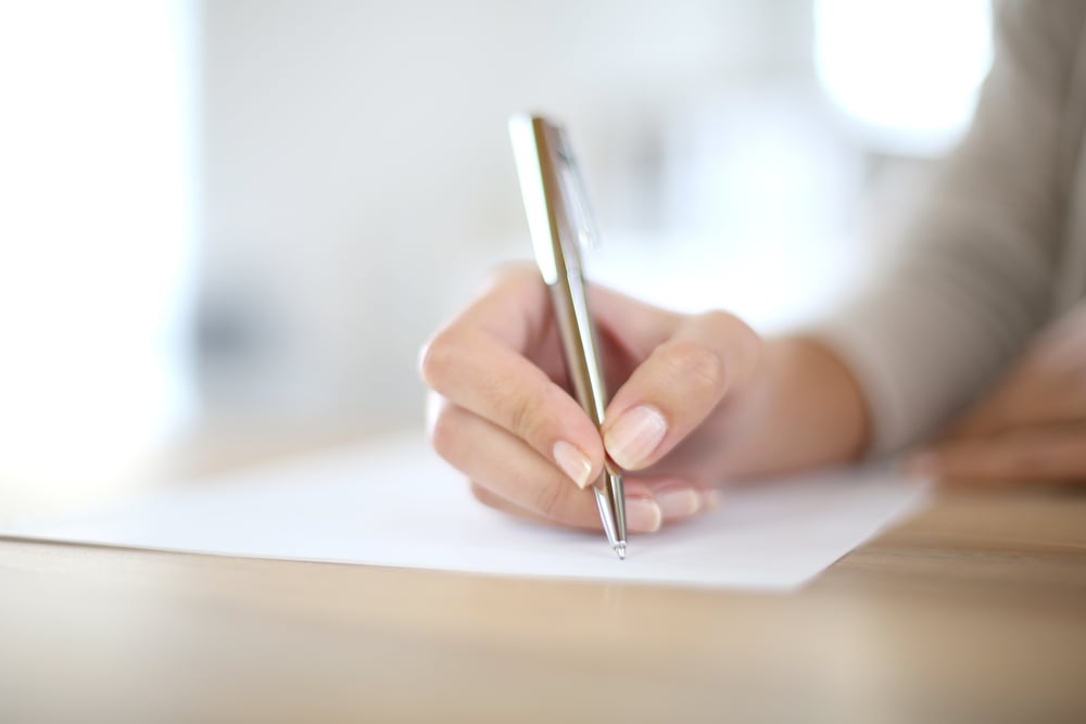 A person's hand holding a pen, writing on a sheet of paper placed on a wooden table.