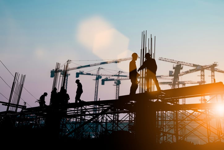 Silhouetted construction workers on a building framework, diligently practicing construction risk management, with cranes in the background during sunset.