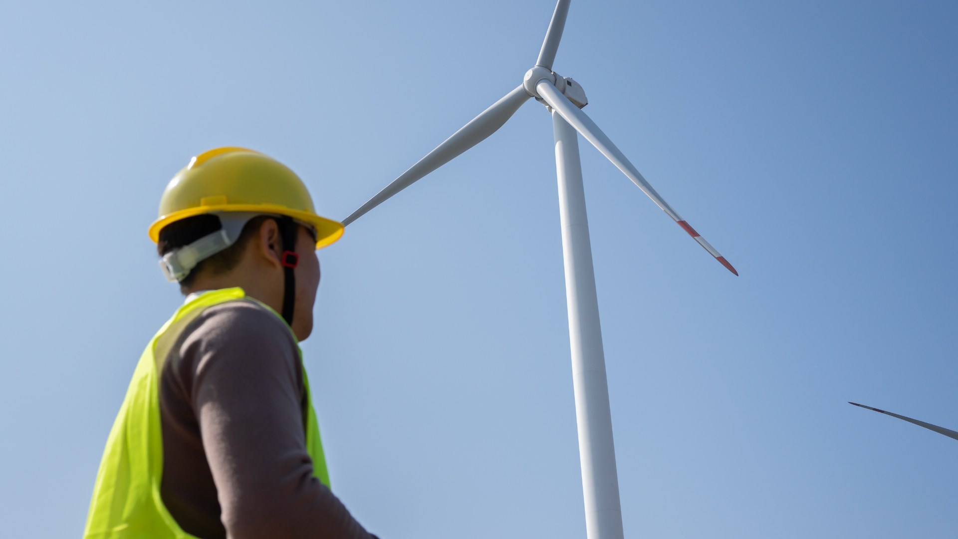 A worker wearing a hard hat and safety vest looks up at a large wind turbine on a clear day