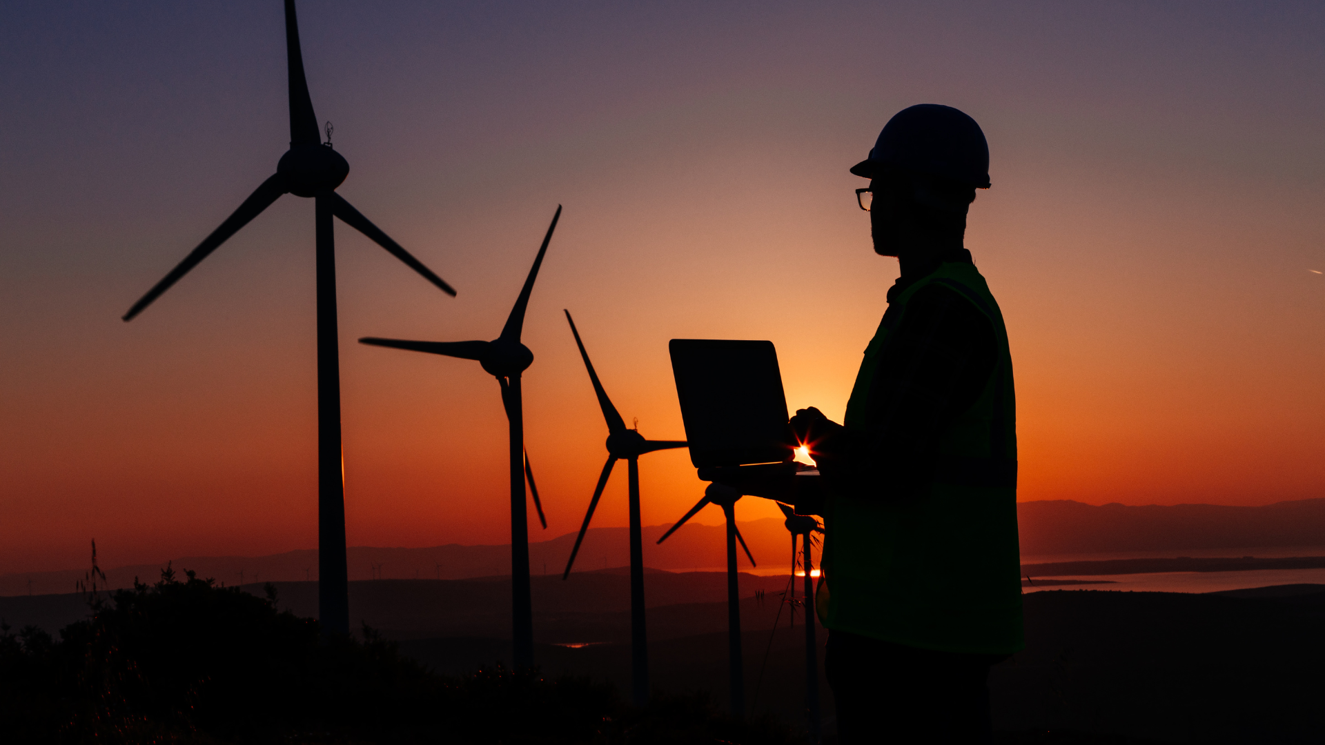 A person in a hard hat and reflective vest uses a laptop near wind turbines at sunset