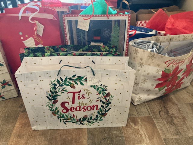 Several holiday-themed gift bags, including one with the text "Tis the Season," are arranged on a wooden floor, containing wrapped presents.