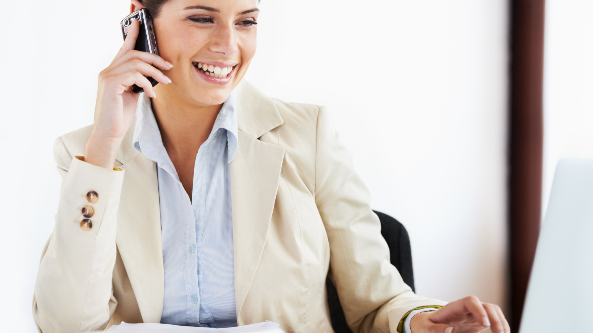 A woman in a beige blazer is smiling while talking on a mobile phone and working on a laptop, embodying the professionalism of an enterprise risk management consultant.