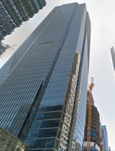 A tall, modern skyscraper with a glass exterior is shown from below. Adjacent is a yellow construction crane