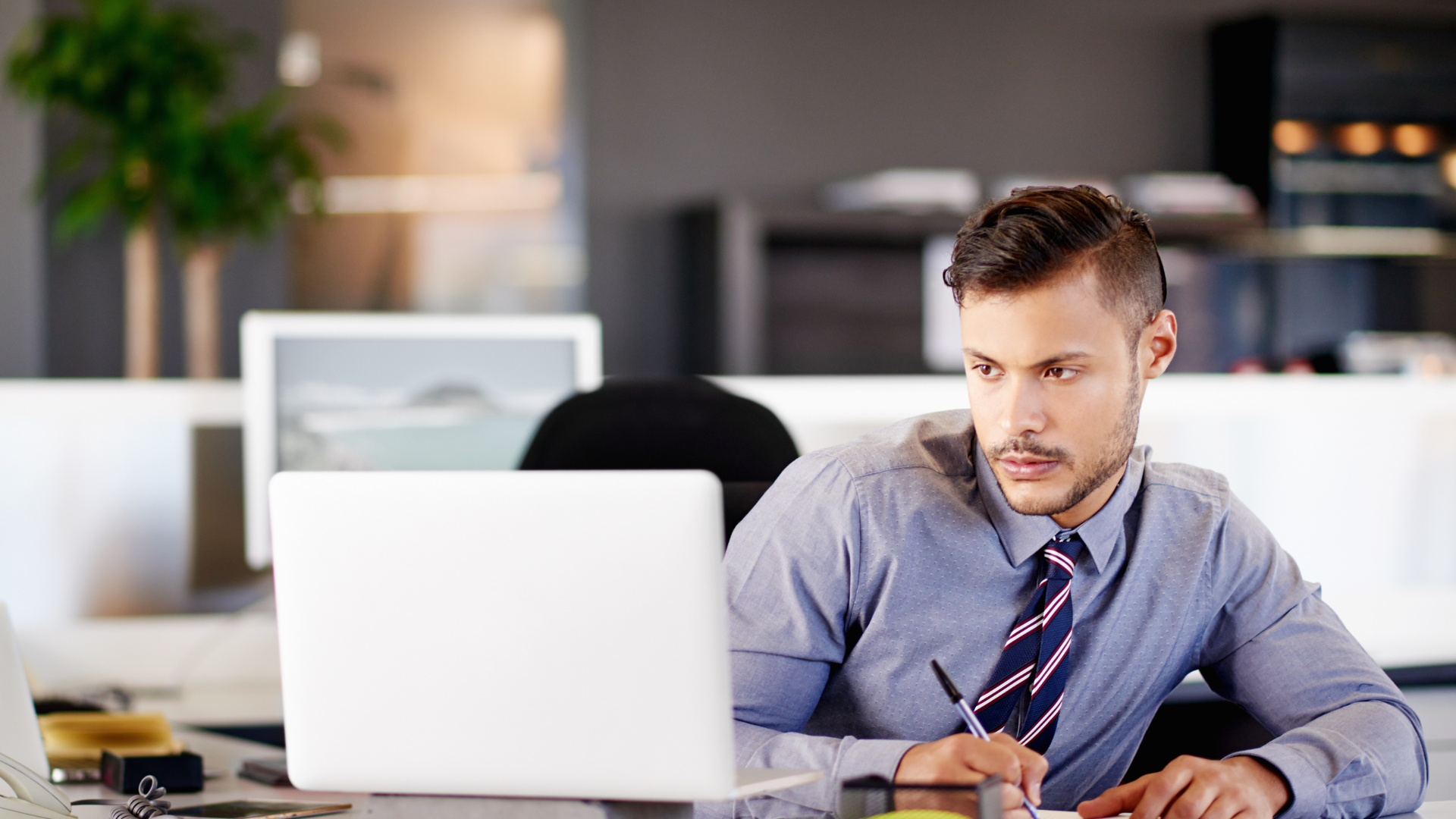 A financial risk management consultant in a shirt and tie sits at a desk, looking at a laptop screen while writing on a notepad.