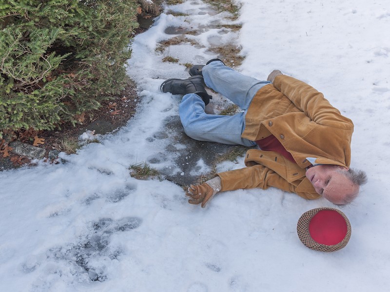 An elderly man wearing a brown coat and gloves has fallen on a snowy path.