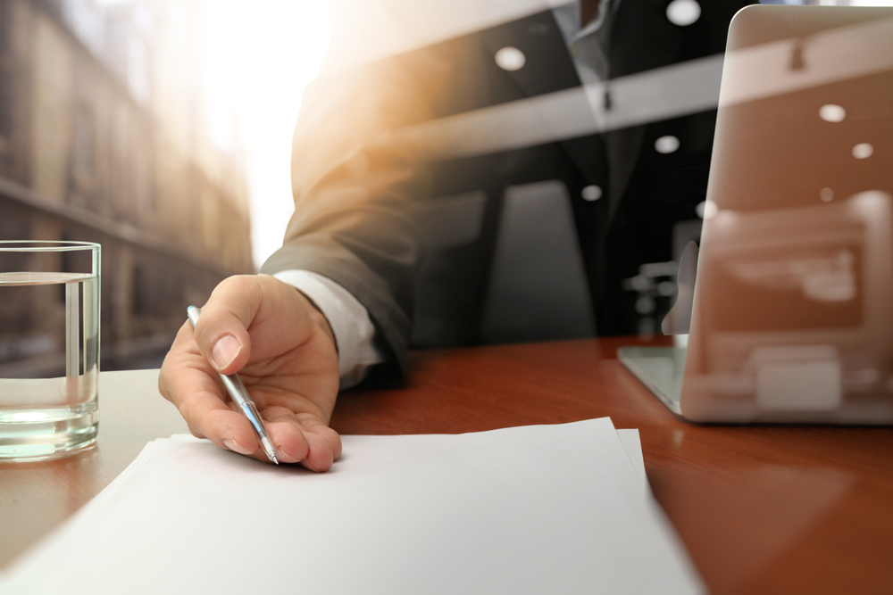 A person in a suit holding a pen over a blank piece of paper at a desk, with a glass of water and a laptop nearby.