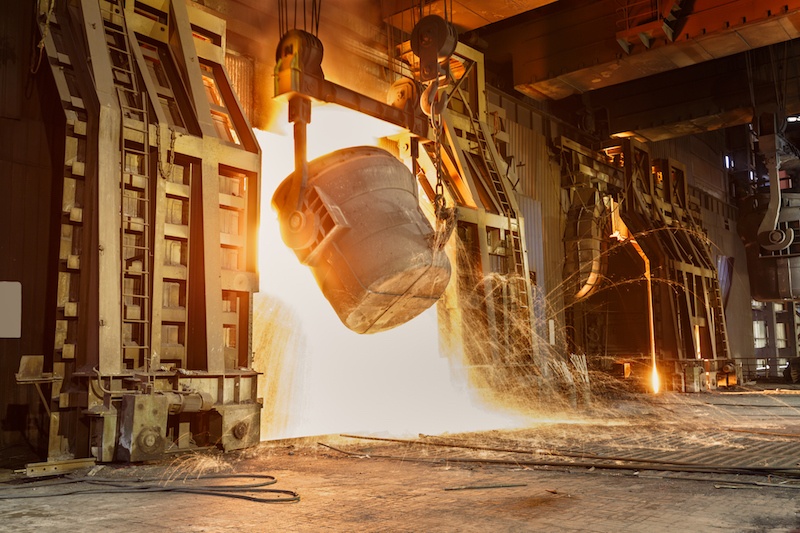 A cauldron of molten metal is being poured out in a factory, emitting bright light and sparks, with heavy machinery in the background.