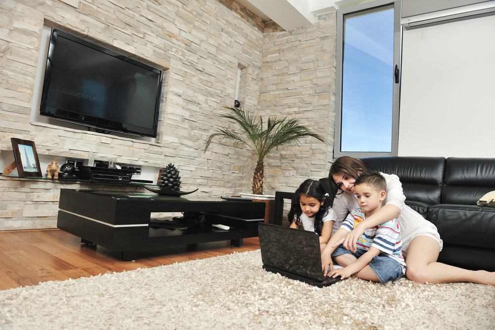 A woman and two children sit on a black couch, looking at a laptop in a modern living room