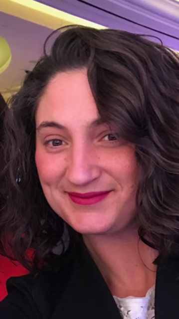 Person with curly dark hair smiling and wearing a black top in a close-up portrait.