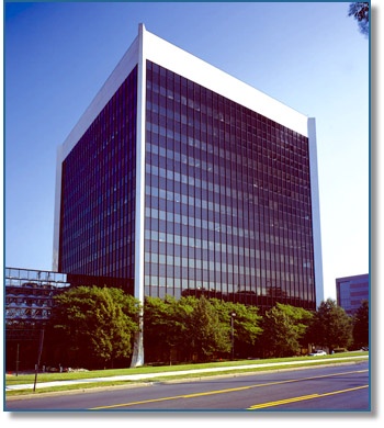 A tall, modern office building with a glass facade under a clear blue sky, surrounded by trees and a road in the foreground.