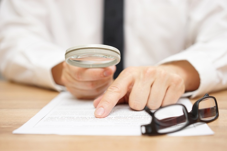 A person in a white shirt uses a magnifying glass to read a document on cyber liability insurance at a desk, with a pair of glasses placed nearby.