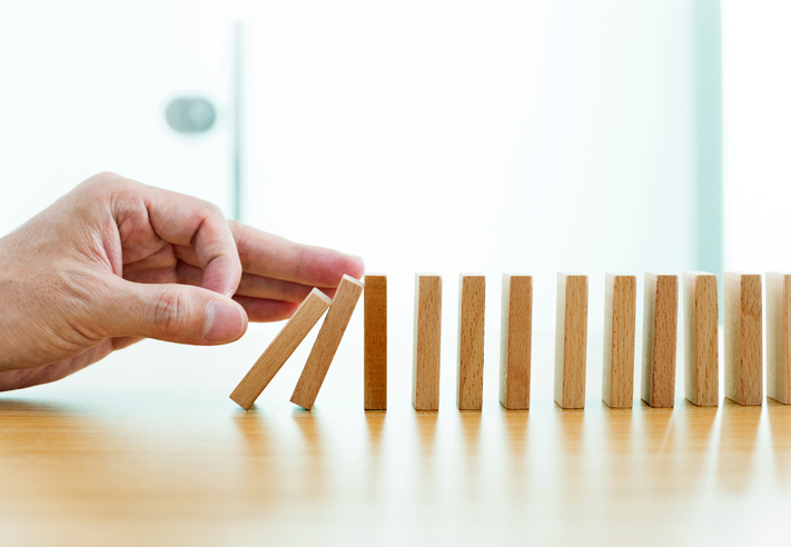 A hand is pushing the first of several wooden dominoes, causing them to fall in sequence on a wooden surface.