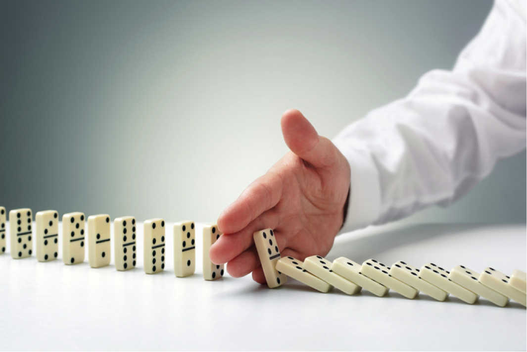 A hand in a white shirt executing risk treatment by stopping a line of falling dominoes on a white surface.