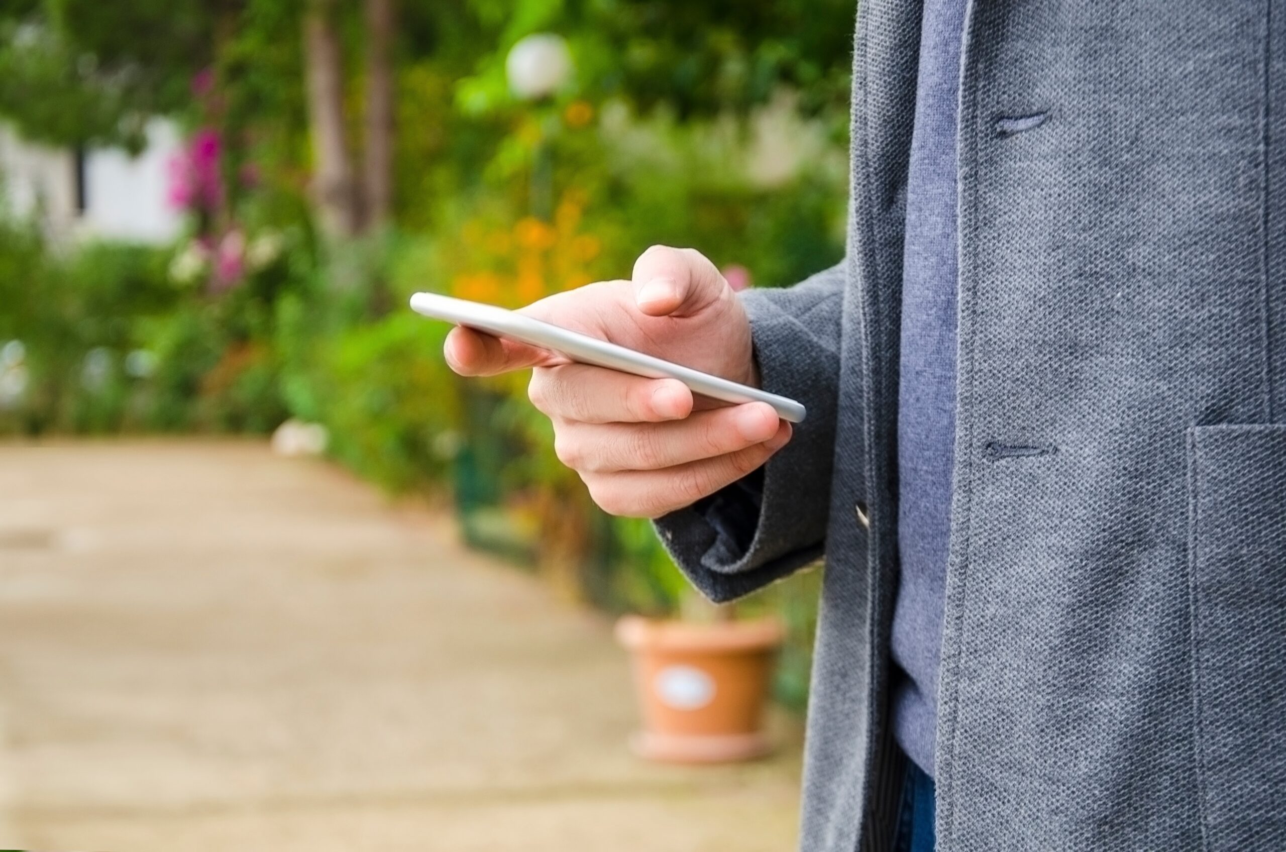 A person wearing a gray coat is holding and looking at a smartphone outdoors with a blurred garden background.