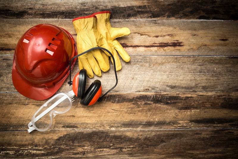 A red safety helmet, yellow gloves, protective eyewear, and ear muffs on a wooden surface.