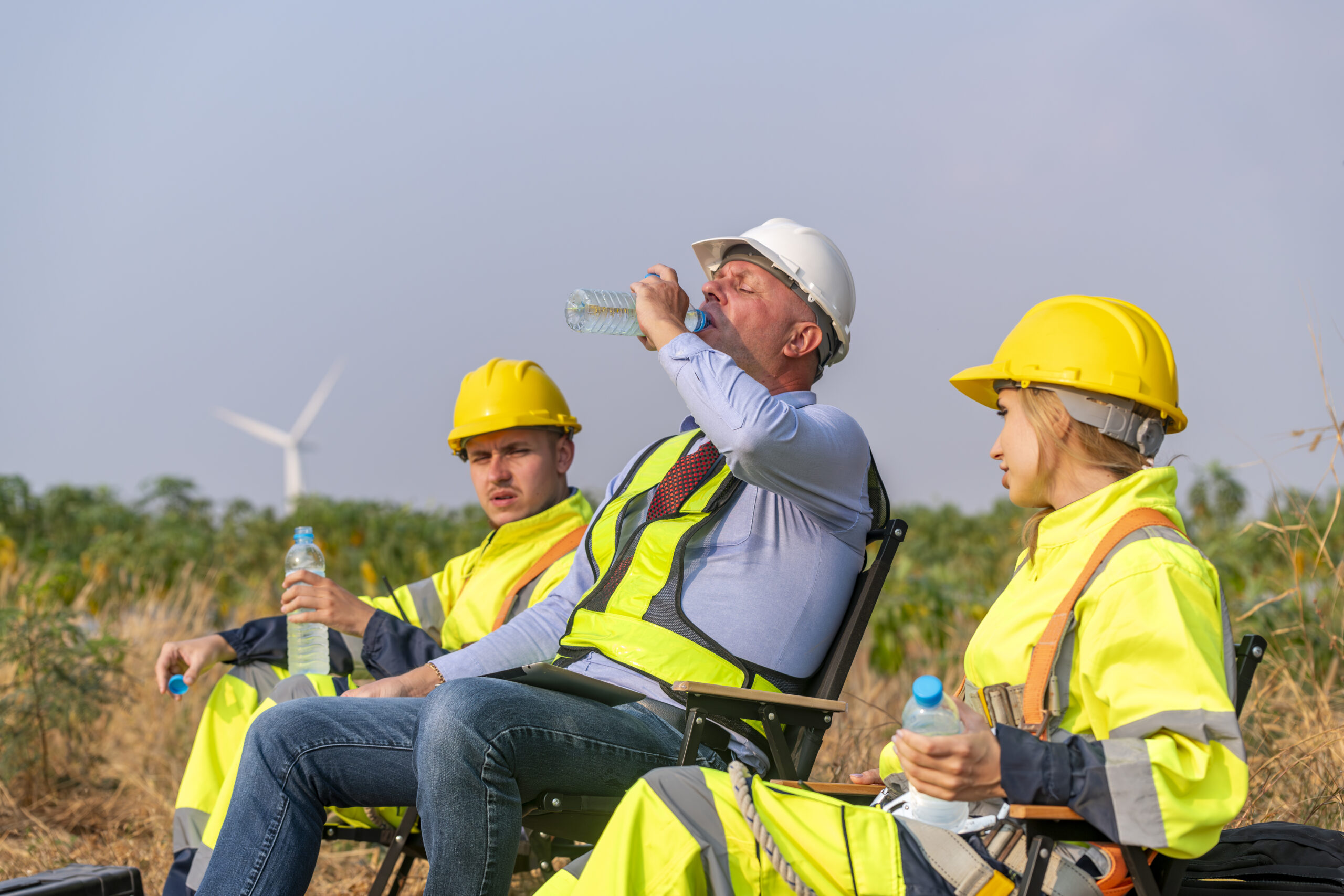 Three construction workers in safety gear sit outdoors on chairs, drinking water to ensure worker safety. In the background, an impressive wind turbine stands tall.